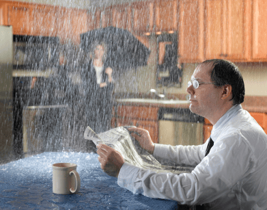 Man in white shirt reading newspaper indoors while heavy rain pours down a window behind him.