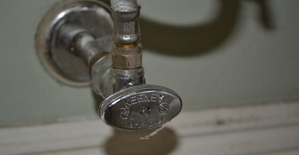 Close-up of an old brass Regency faucet handle with decorative knurled details and mounting hardware on a white wall.