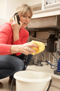 Woman in red shirt crouched by dishwasher on phone, holding yellow sponge over bucket catching water leak.