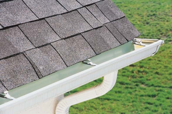 White gutter system attached to gray asphalt shingle roof with downspout visible above green lawn.
