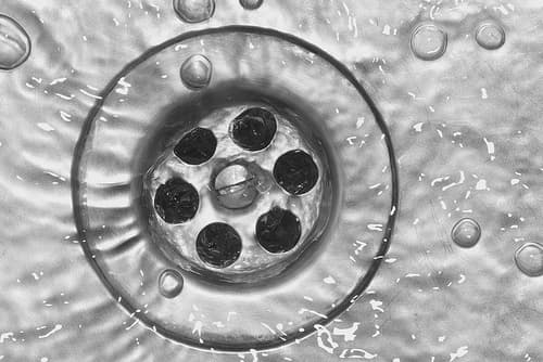 Close-up view of a drain opening surrounded by water droplets and bubbles on a wet surface.