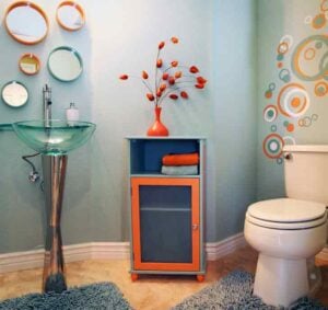 Contemporary bathroom featuring a glass pedestal sink, orange and blue storage cabinet with decorative flowers, colorful circular wall decals, and white toilet.