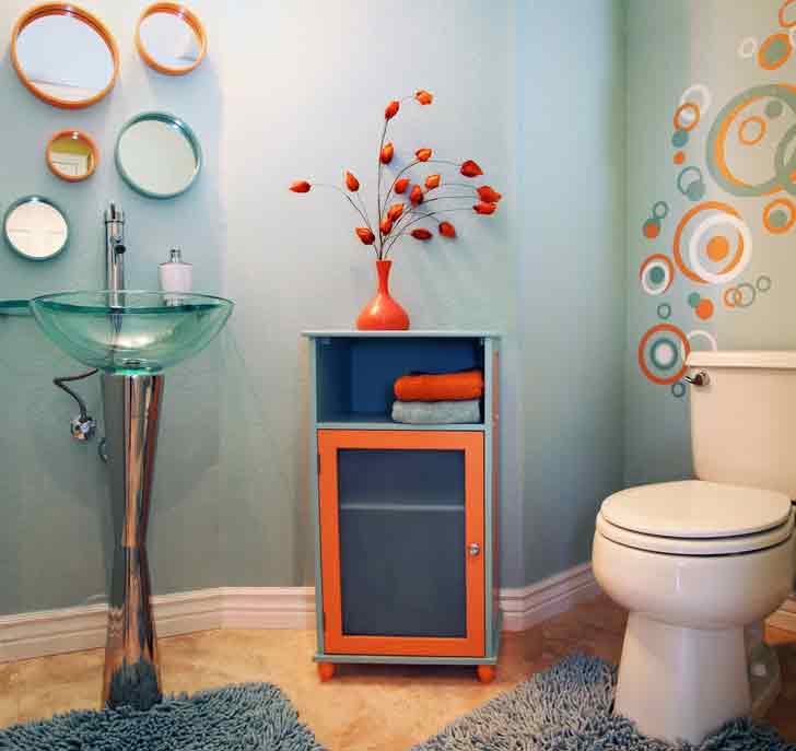Contemporary bathroom featuring a glass pedestal sink, orange and blue storage cabinet with decorative flowers, colorful circular wall decals, and white toilet.