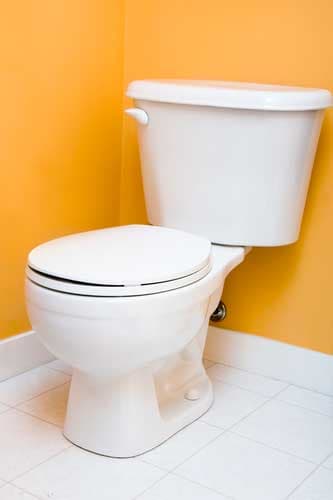 White ceramic toilet with closed seat lid and matching tank against a bright yellow bathroom wall with white tile flooring.