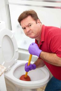 Man in red shirt wearing purple gloves uses a plunger to unclog a white toilet bowl.