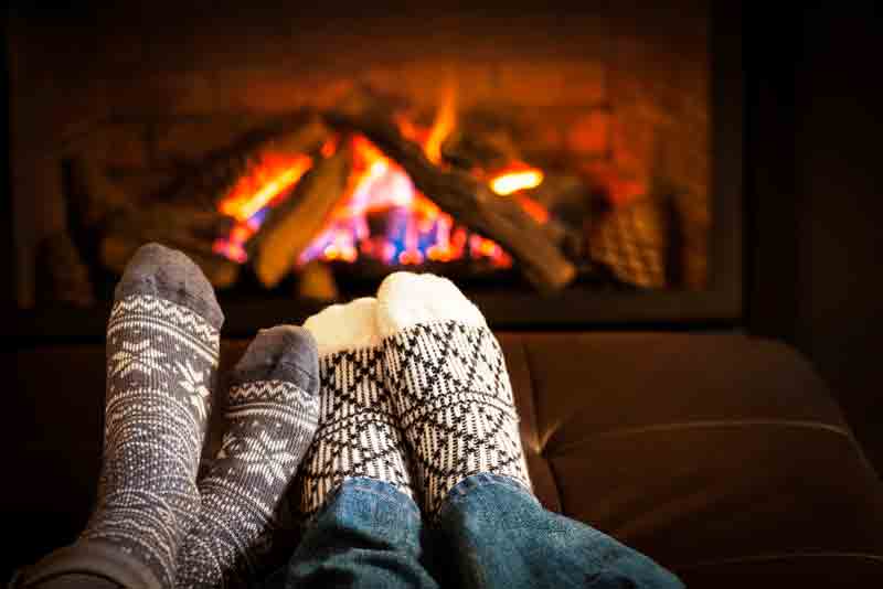 Person wearing warm socks relaxing with feet up near a fireplace with glowing flames and logs.