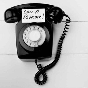 Black vintage rotary telephone with handset on white background, displaying "Call A Plumber!" message on note card.