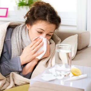 Woman with cold or flu sitting on couch holding tissue to her nose with water glass and tissues nearby.