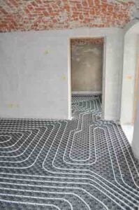 Underfloor heating pipes laid in curved patterns on concrete floor in empty room with exposed brick ceiling.