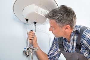 Male technician installing or servicing a ceiling-mounted water heater tank against white wall.