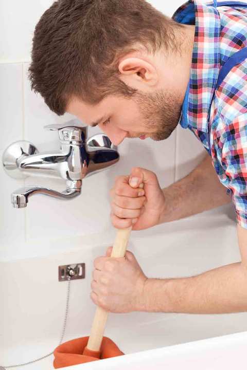 Man in plaid shirt using a plunger to clear a clogged bathroom sink drain at a white wall-mounted faucet.