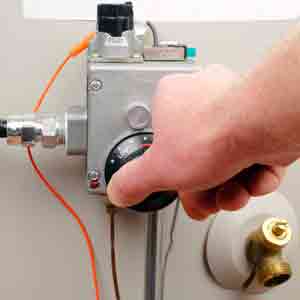 Technician's hand adjusting thermostat dial on gas water heater with orange tubing and brass valve connections.