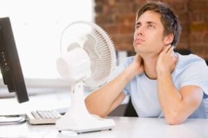 Man in light blue shirt sitting at desk with computer monitor and white fan, hand on neck appearing uncomfortable or hot.