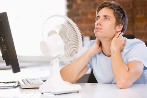 Man in light blue shirt sitting at desk with computer monitor and white fan, hand on neck appearing uncomfortable or hot.