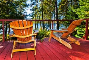 Two orange Adirondack chairs on a red wooden deck with a lake and evergreen trees visible in the background.