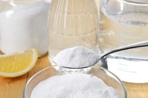 Baking soda being poured into a measuring spoon over a sink with lemon, vinegar, and cleaning supplies nearby.