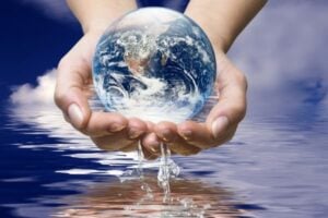 Hands cupping a transparent globe showing Earth with water dripping below against a blue sky background.