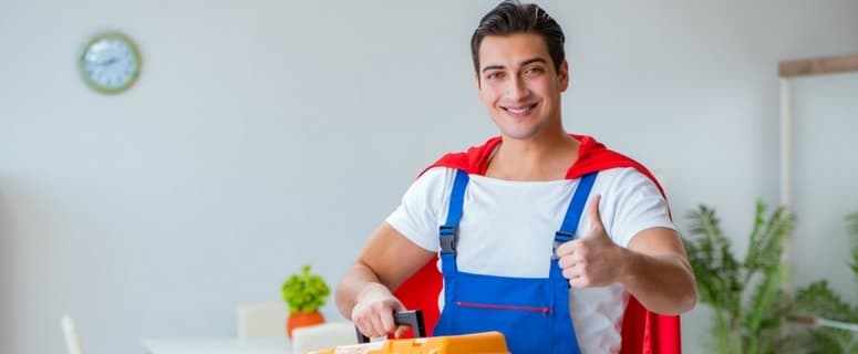 Young man in blue work overalls and red cape gives thumbs up while holding a toolbox indoors.