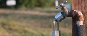 Weathered outdoor water faucet mounted on wooden post with blue handle and metal padlock attached.