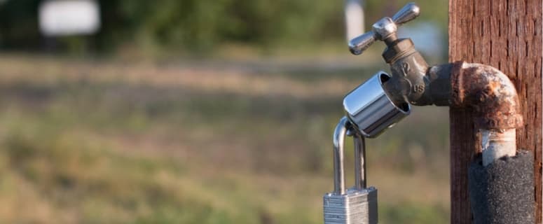 Weathered outdoor water faucet mounted on wooden post with blue handle and metal padlock attached.