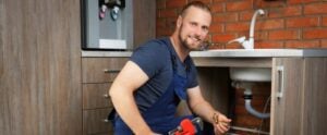 Male plumber in blue shirt working on under-sink plumbing replacement with tools and red pipe wrench visible.