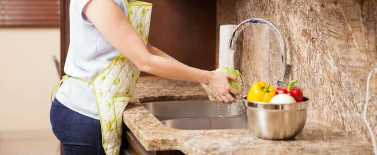 Woman in apron washing fresh vegetables at a granite countertop sink with a metal colander of produce nearby.