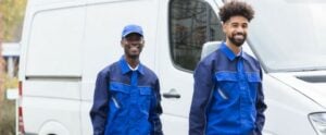 Two smiling male HVAC technicians in blue work uniforms and caps standing in front of a white service van.