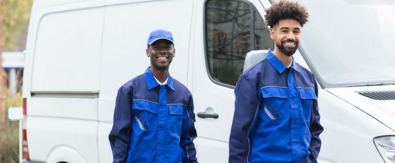 Two smiling male HVAC technicians in blue work uniforms and caps standing in front of a white service van.