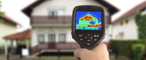 Handheld thermal imaging camera displaying colorful heat map of residential house showing temperature distribution across building facade.