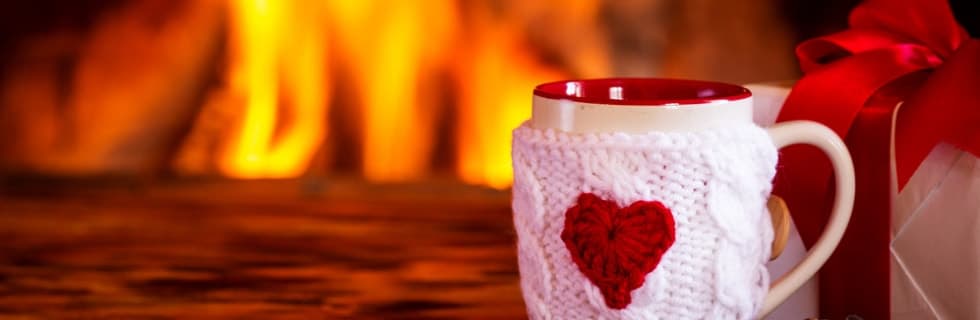 Pink knit mug cozy with red heart design sits by a glowing fireplace with gift boxes nearby.