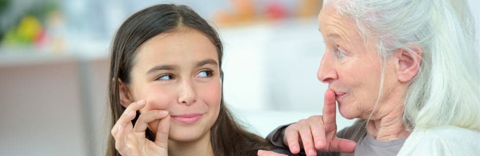 An older woman with gray hair speaks to a young girl about skincare, both looking at the girl's face with concern about skin texture.