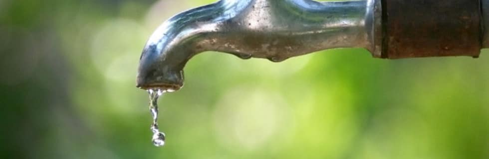 Close-up of a metal outdoor faucet with water droplets dripping from the spout against a blurred green background.