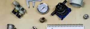 Assorted plumbing supplies including pressure gauge, valve components, calipers, and brass fittings arranged on tan surface.