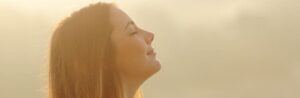 Woman with eyes closed, face tilted upward, breathing fresh air during golden hour sunlight.