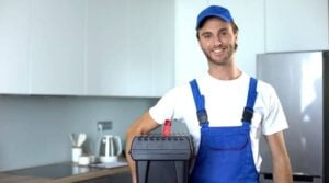 Smiling plumber in blue uniform and cap holding a black toolbox in a modern kitchen.