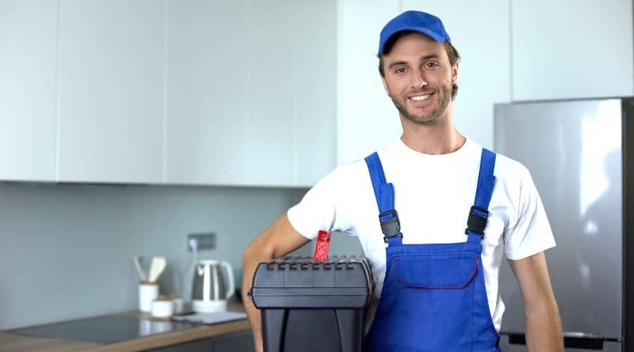 Smiling plumber in blue uniform and cap holding a black toolbox in a modern kitchen.