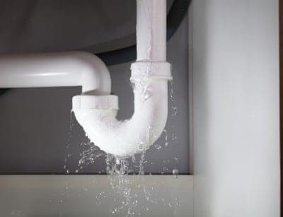 Water dripping from a white PVC pipe elbow joint under a sink, showing active plumbing leak onto surface below.
