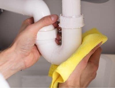 Hand holding white P-trap pipe with yellow cleaning cloth, removing debris and buildup from plumbing fixture.