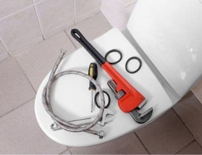 Plumbing tools laid out on a white toilet seat including a red pipe wrench, flexible hose connector, and metal rings on a tiled bathroom floor.