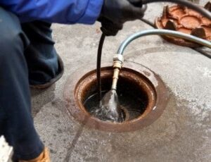 Plumber using a drain snake tool to clear a clogged floor drain with water flowing through the copper fitting.
