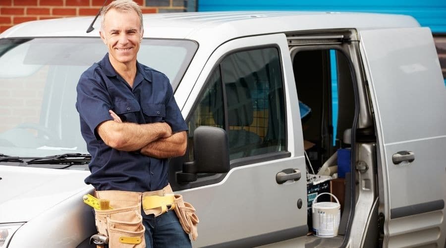 Smiling male tradesman in navy work shirt and tool belt stands with arms crossed beside white service van with open door.