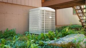 White air conditioning condenser unit installed against a brown building wall surrounded by green plants and landscaping.