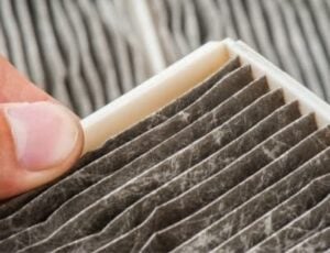 A finger pointing to a clogged gray air filter showing accumulated dust and debris buildup compared to a clean white filter section.