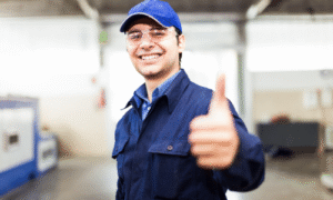 Smiling technician in blue uniform and cap giving thumbs up gesture in workshop setting.