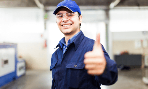Smiling technician in blue uniform and cap giving thumbs up gesture in workshop setting.