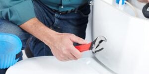 Plumber in blue shirt holding adjustable wrench while working on white toilet installation or repair.