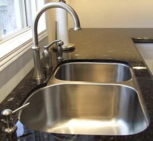 Modern double basin stainless steel kitchen sink with curved chrome faucet and soap dispenser on dark countertop.