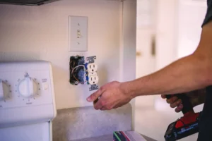 Electrician's hand installing a white duplex outlet into a wall box with exposed blue and black wires, light switch visible above.
