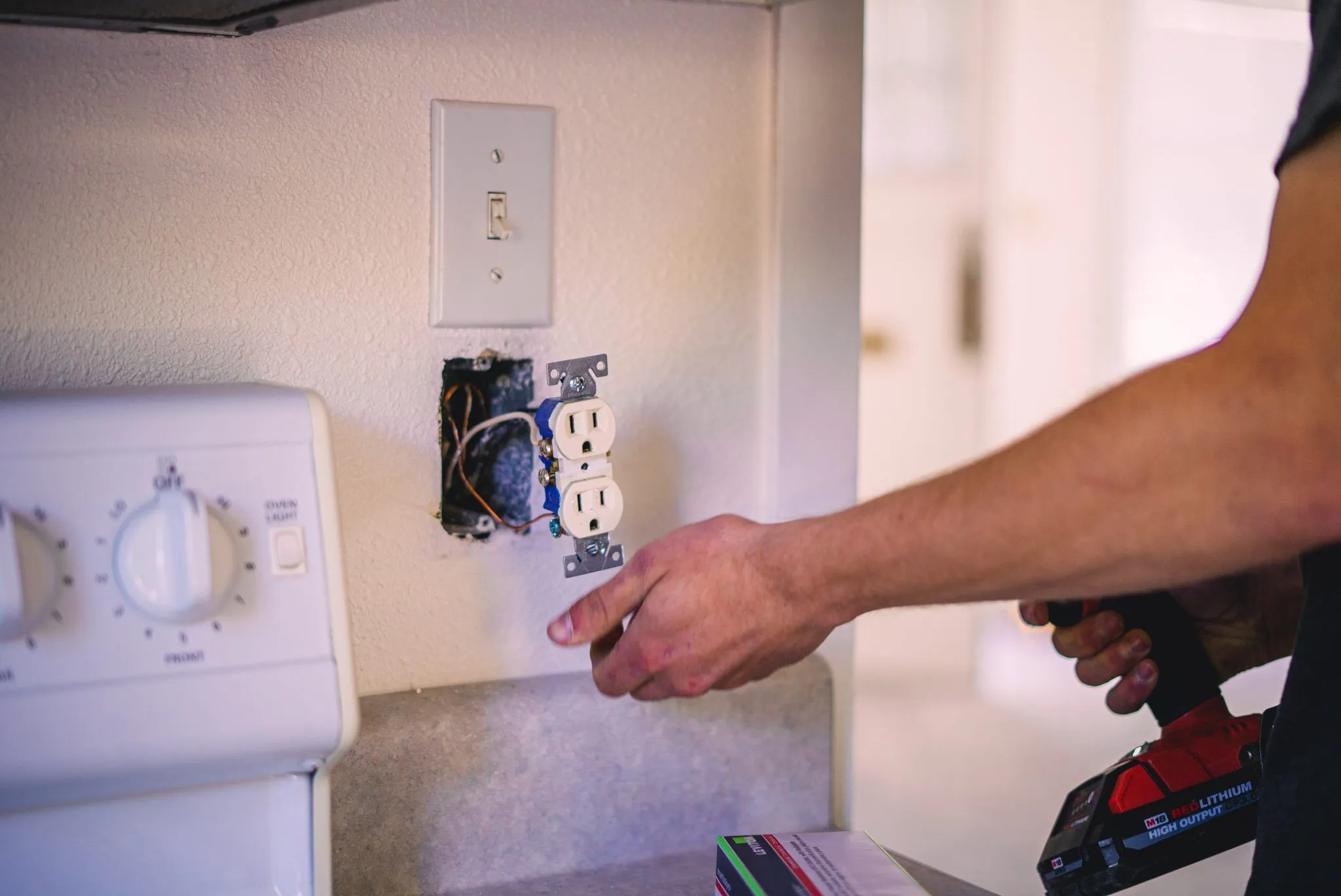Electrician's hand installing a white duplex outlet into a wall box with exposed blue and black wires, light switch visible above.
