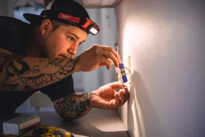 Tattooed electrician wearing Milwaukee headlamp installs electrical outlet into wall during construction work.